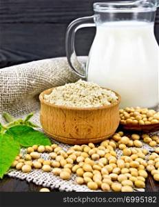 Soy flour in the bowl, soybeans in a spoon and on a napkin of burlap, milk in a jug, soya leaf against the background of a dark wooden board