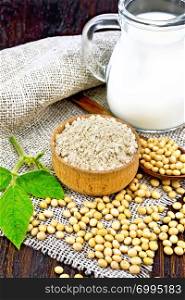 Soy flour in the bowl, soybeans in a spoon and on sackcloth, milk in a jug, green soya leaf against a dark wooden board