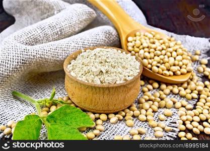 Soy flour in the bowl, soybeans in a spoon and on sackcloth, green soya leaf on a wooden board background