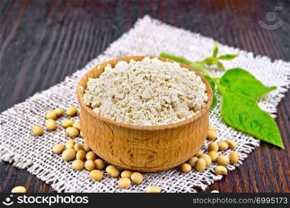Soy flour in the bowl, soybeans on burlap, green leaf on a wooden board background