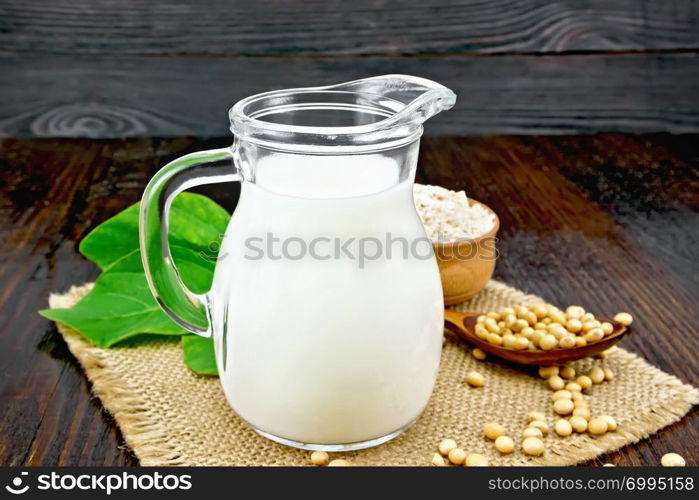 Soy milk in a jug, flour in a bowl and a fresh green leaf, soybeans in a spoon and burlap on the background of a dark wooden board