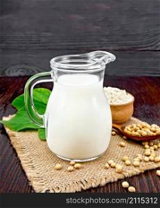 Soy milk in a jug, flour in a bowl and green leaf, soybeans in a spoon and burlap on the background of a dark wooden board