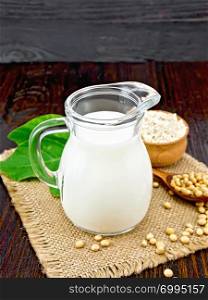 Soy milk in a jug, flour in a bowl and green leaf, soybeans in a spoon and burlap on a wooden board background