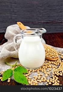 Soy milk in a jug, fresh green leaf, soybeans in a spoon and burlap on a wooden board background