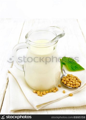 Soy milk in a jug, green leaf, soybeans in a spoon on a napkin on the background of wooden boards