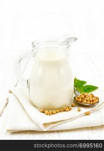 Soy milk in a jug, green leaf, soybeans in a spoon on a napkin against the background of a light wooden board