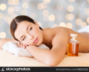 spa, beauty, people and body care concept - happy woman lying on the massage desk with oil bottle over holidays lights background