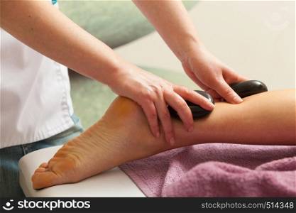 Spa relaxation, healthy pleasure concept. Woman lying on stomach, female masseuse doing legs and feet massage with hot stones. Masseuse doing legs massage with hot stones