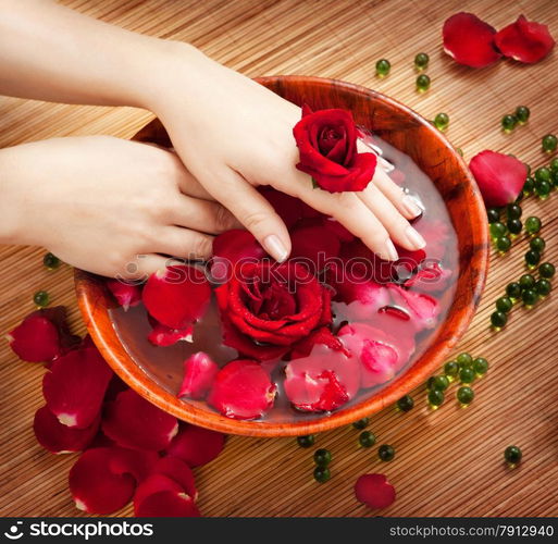 Spa Salon: Beautiful Female Hands with French Manicure in the Bamboo Bowl of Water with Red Roses and Rose Petals on the Straw Mat