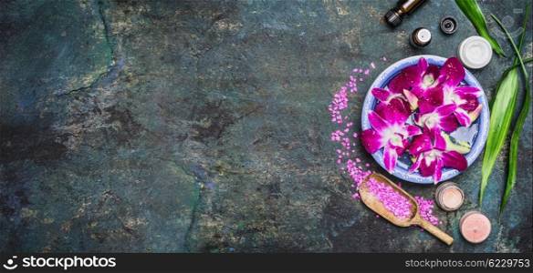 Spa setting with water bowls , pink orchid flowers ,sea salt, cosmetic cream and essential oil on dark rustic background , top view, place for text, horizontal, banner. Wellness concept