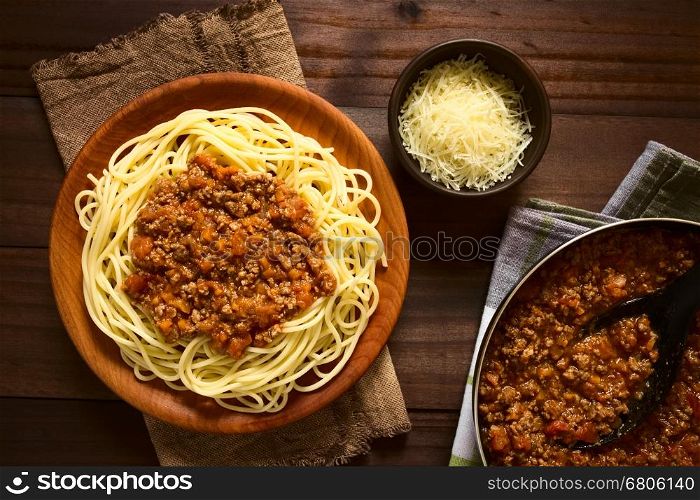 Spaghetti with homemade bolognese sauce made of fresh tomato, mincemeat, onion, garlic and carrot, served on wooden plate with grated cheese and skillet with sauce on the side, photographed overhead with natural light