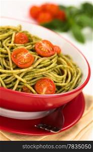 Spaghetti with pesto and baked cherry tomato halves served in a red bowl with a fork beside on white wood, with cherry tomatoes and basil leaf in the back (Selective Focus, Focus one third into the dish). Spaghetti with Pesto and Tomato
