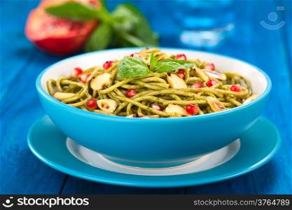 Spaghetti with pesto, pomegranate and roasted almond halves garnished with a basil leaf and served in a blue bowl on blue wood, with glass of water, half pomegranate and basil leaf in the back (Selective Focus, Focus on the front of the basil leaf on the dish). Spaghetti with Pesto, Pomegranate and Almonds