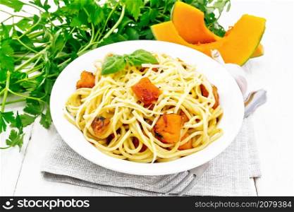 Spaghetti with pumpkin, onion and garlic in a plate on a towel, basil, parsley and fork on the background of light wooden board