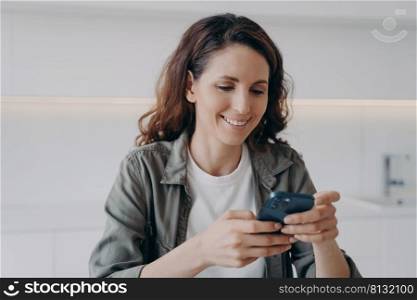 Spanish girl in casual wear is typing and receiving messages. Young happy woman is chatting on smartphone at home. Leisure and communication concept. Homey girl having good time.. Spanish girl in casual wear is typing and receiving messages. Happy woman with smartphone at home.