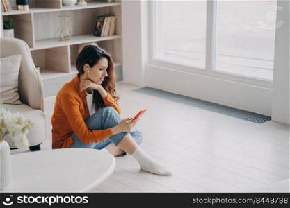 Spanish girl is sitting on floor at home near window in morning and listening to music on telephone. Trendy young woman in airpods. Downloading tracks to phone. App for online music listening.. Spanish girl is sitting on floor at home near window in morning and listening to music on telephone.