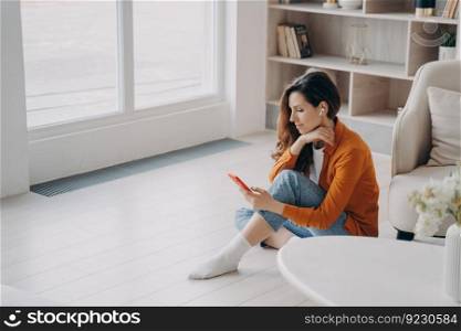 Spanish girl is sitting on floor at home near window in morning and listening to music on telephone. Trendy young woman in airpods. Downloading tracks to phone. App for online music listening.. Spanish girl is sitting on floor at home near window in morning and listening to music on telephone.