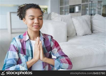 Spanish young woman is sitting on floor in lotus position with her eyes closed. Teenage afro girl is practicing yoga at home. Tranquility and meditation. Stress relief and mental health concept.. Spanish girl is sitting in lotus position with her eyes closed. Stress relief and mental health.