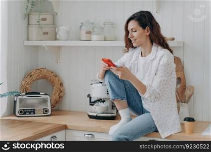 Spanish young woman typing on smartphone. Happy relaxed girl with telephone sitting on worktop of table at kitchen alone. Modern luxurious scandinavian interior. Lifestyle concept.. Spanish woman typing on smartphone. Girl sitting on worktop of table at kitchen alone.