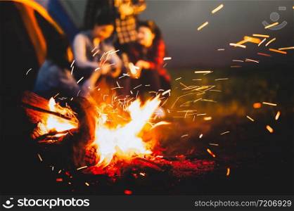 Sparking bonfire with tourist people sit around bright bonfire near camping tent in forest in summer night background. Group of student at outdoor fire fuel. Travel activity and long vacation weekend