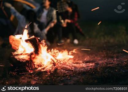Sparking bonfire with tourist people sit around bright bonfire near camping tent in forest in summer night background. Group of student at outdoor fire fuel. Travel activity and long vacation weekend
