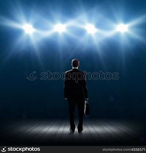 Speaker on stage. Rear view of businessman standing in lights of stage