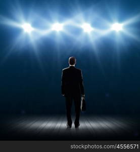 Speaker on stage. Rear view of businessman standing in lights of stage