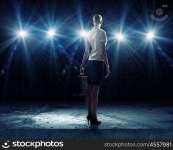 Speaker on stage. Rear view of businesswoman standing in lights of stage