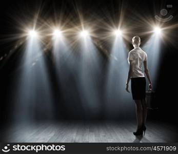 Speaker on stage. Rear view of businesswoman standing in lights of stage