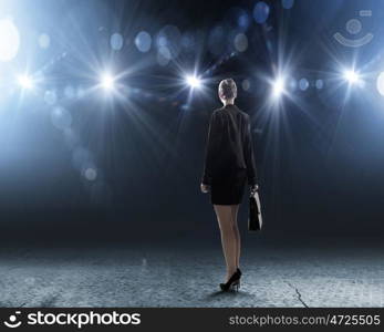 Speaker on stage. Rear view of businesswoman standing in lights of stage