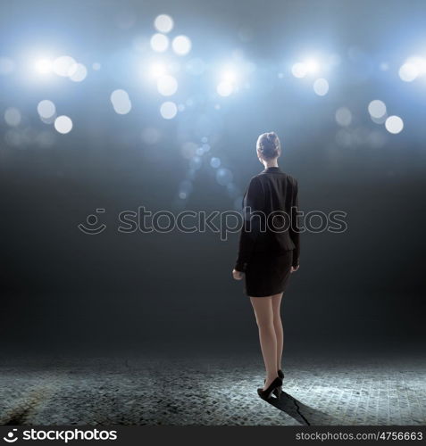 Speaker on stage. Rear view of businesswoman standing in lights of stage