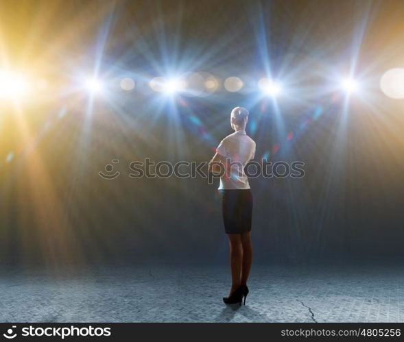 Speaker on stage. Rear view of businesswoman standing in lights of stage