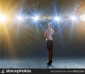 Speaker on stage. Rear view of businesswoman standing in lights of stage