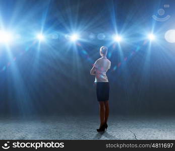 Speaker on stage. Rear view of businesswoman standing in lights of stage