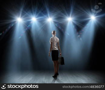 Speaker on stage. Rear view of businesswoman standing in lights of stage