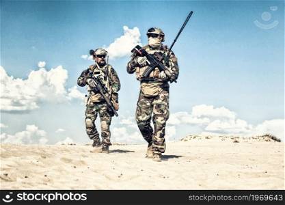Special operations command soldiers, elite forces fighters, marine corps riders team in woodland camo uniform, equipped satellite radio system, patrolling sandy area with service rifles in hands. Armed commando soldiers walking in sandy area