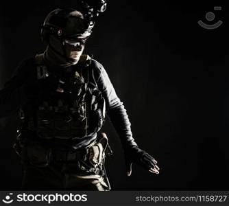 Special operations fighter in helmet with night-vision, thermal imaging device, load carrier carefully moving with caution in darkness, holding hand on pistol, ready for fight during dangerous mission. Army infantryman moving in darkness with caution