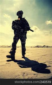 Special operations forces soldier in camouflage combat uniform, helmet and glasses walking in desert with service rifle in hands low angle view. War conflict, military campaign in Middle East region. Equipped army soldier with rifle walks in desert