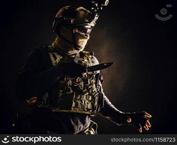 Special operations soldier, commando fighter, modern warfare combatant in combat uniform, helmet with night-vision, wearing mask and glasses, sneaking in darkness for sentry quiet removal with knife. Commando fighter creeping in darkness with knife