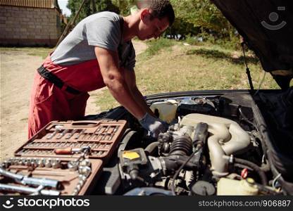 Specialist auto mechanic in the car service, checks the car, engine, engine, carburetor. Concept: repair of machines, fault diagnosis, repair specialist, technical maintenance. Mechanic standing in front of the open hood and repairing the car
