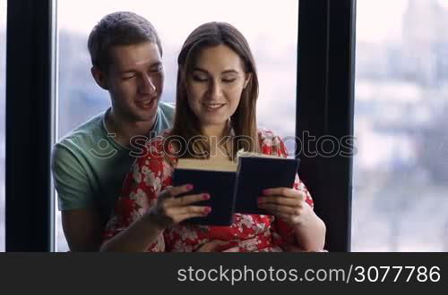 Spending nice time at home. Beautiful young loving couple bonding to each other and smiling while woman reading a book agianst wide window background. Handsome man cuddling attractive woman from behind while relaxing in modern apartment.