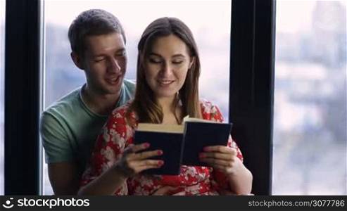 Spending nice time at home. Beautiful young loving couple bonding to each other and smiling while woman reading a book agianst wide window background. Handsome man cuddling attractive woman from behind while relaxing in modern apartment.