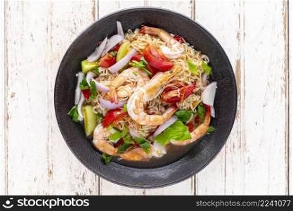 spicy and sour instant noodle salad with shrimp, minced pork, tomato, lime, red onion, shallot and celery in black ceramic plate on white old wood texture background, top view, Thai food, Yum Mama