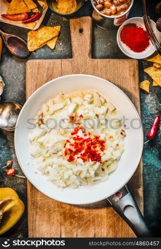 Spicy food cooking preparation. Frying pan with cut onion, garlic and chili on rustic cutting board with spices ingredients, top view. Mexican cuisine