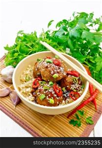 Spicy Korean meatballs in sauce of starch, soy sauce, vinegar and apricot jam, sprinkled with green onions, hot peppers and sesame seeds in a bowl on a bamboo napkin against wooden board background