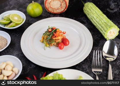 Spicy lime pork salad with bitter melon, galangal, chili, tomatoes and garlic in a white plate on a black cement floor.