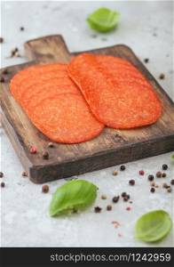 Spicy pepperoni salami with basil and pepper on wooden chopping board and light background.