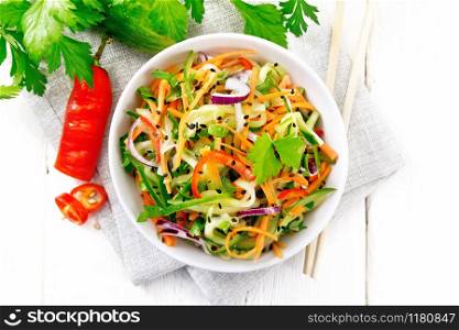 Spicy salad of cucumbers, carrots, chili peppers, purple onions, cilantro and black sesame, seasoned with vinegar and lemon juice in a bowl on a towel on wooden board background from above