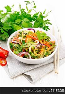 Spicy salad of cucumbers, carrots, chili peppers, purple onions, cilantro and black sesame seeds, seasoned with vinegar and lemon juice in a bowl on a towel on white wooden board background