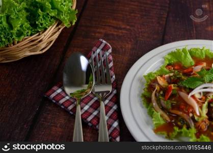 Spicy salad of sardine with tomato sauce mixed with herb in white dish and lettuce leaf in weave basket nearly, the sparken stainless spoon and fork over napkin on wooden table, copy space